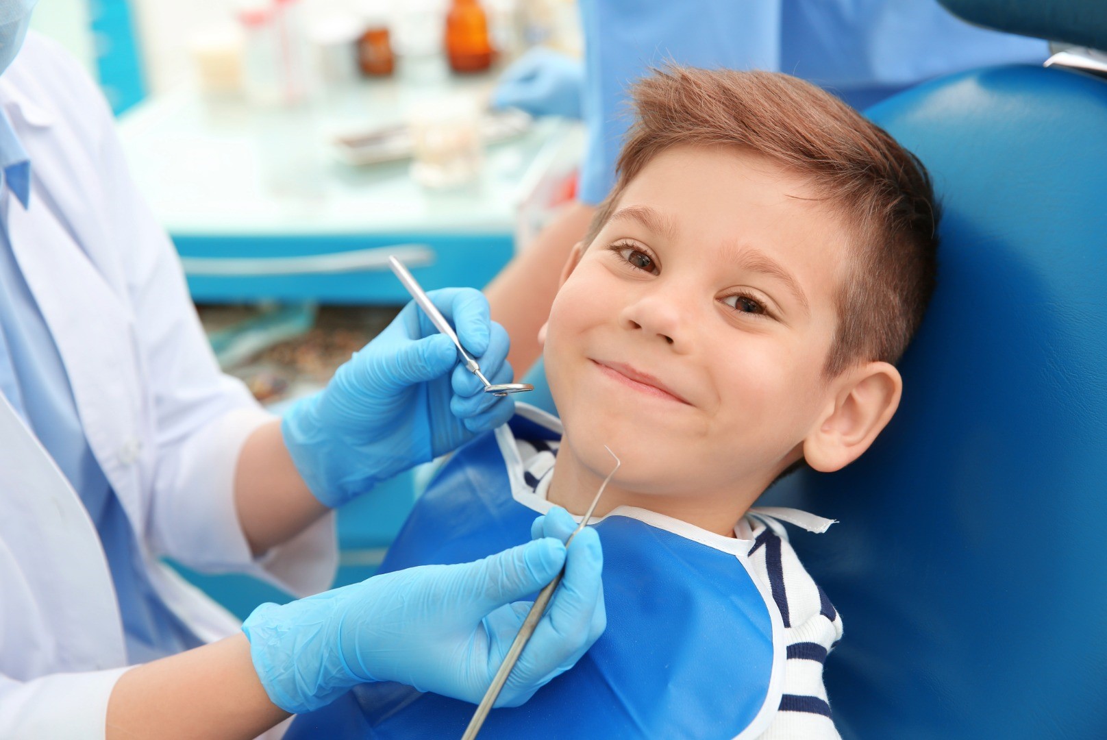 Boy smiles and looks at the camera while sitting on the dentist chairs. A dentist wearing blue gloves and holding dental tools is about to examine the boy's teeth.