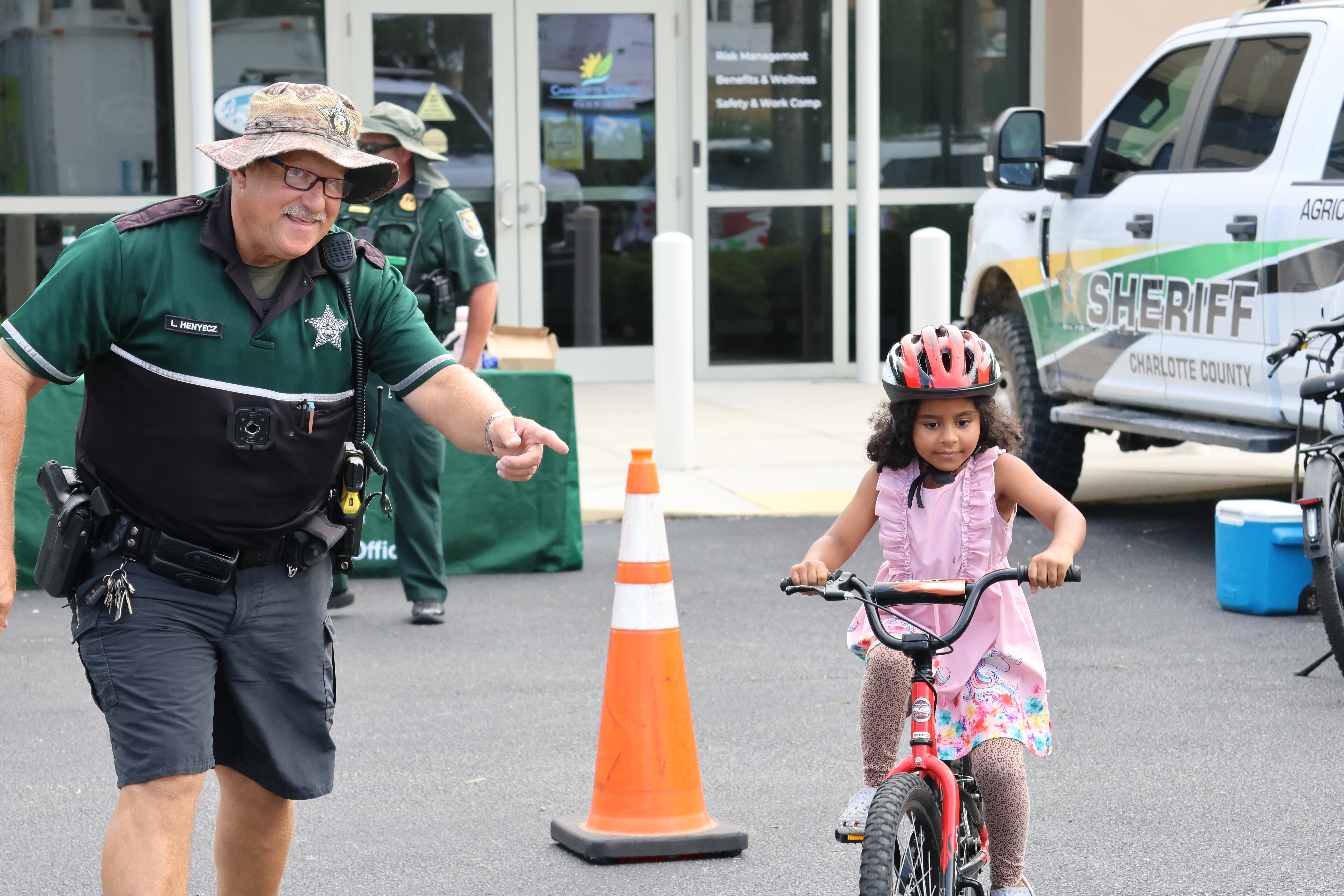 Charlotte County Sheriff's Office deputy jogs next to girl wearing a helmet and riding a bike during the 2025 Back to School Bash at the Florida Department of Health in Charlotte County.